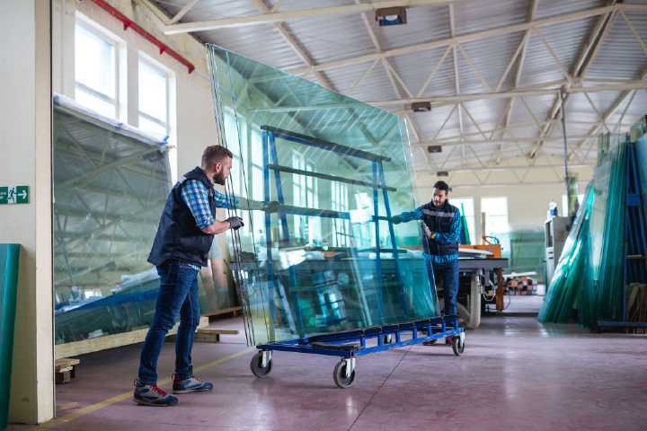 Two individuals in factory environment handling large transparent glass sheets using a blue wheeled cart wearing work attire and gloves with stacked glass panels machinery and high ceiling industrial space in background symbolizing careful handling safety and precision in glass manufacturing.