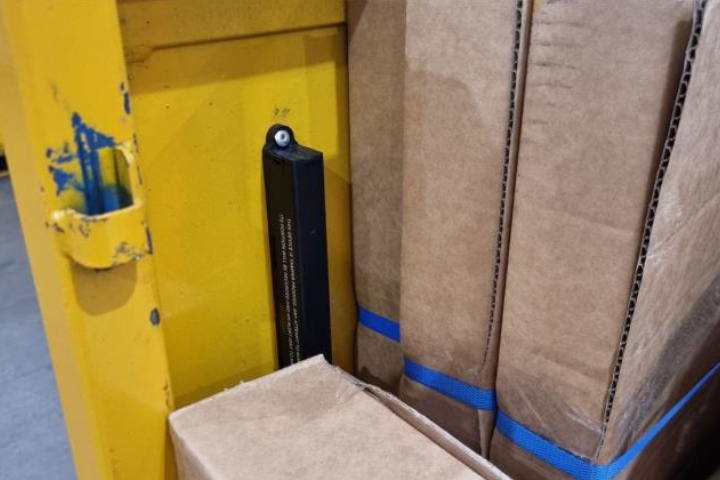 Close up view of warehouse setting with yellow metal structure showing signs of wear fitted with a black rectangular sensor and small white component mounted on it large cardboard boxes secured with blue plastic straps tightly packed together on the right symbolizing industrial safety equipment packaging and storage practices.