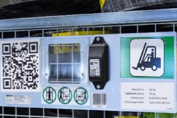 Metal panel attached to wire mesh structure containing a large QR code transparent grid section black electronic component with label pictogram of a forklift three circular safety icons a barcode and a specification label in Swedish indicating weight limits and dimensions for industrial shelving equipment.